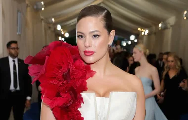 A woman in a strapless dress with a large red floral accessory on the shoulder, posing at a formal event.