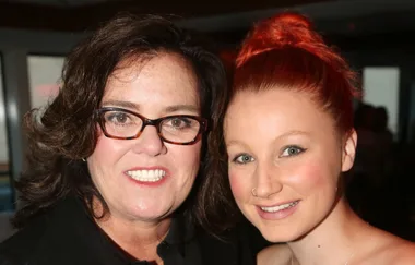Two people smiling at the camera, one with black glasses and dark hair, the other with red hair in a bun.