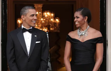 Two individuals in formal attire smiling at each other in a warmly lit room with chandeliers.