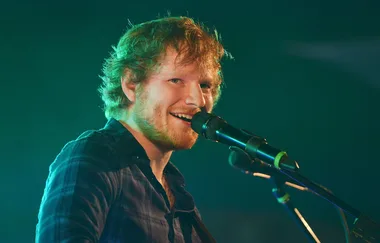 A person with a beard is singing into a microphone on stage, looking towards the camera with a smile.