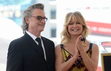 Two people smiling at an event; one in a black suit, the other in a floral dress, hands clasped.