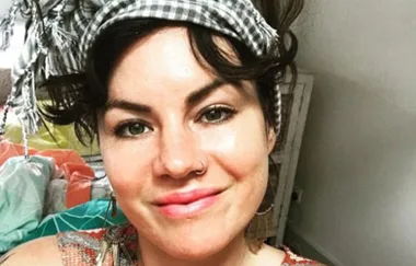 Young woman with a plaid headscarf and a nose ring smiles at the camera, indoors.