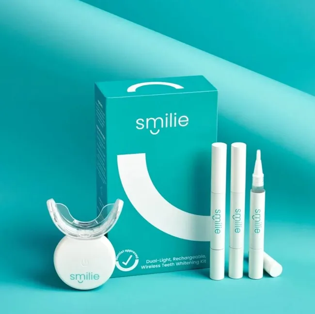 Smilie teeth whitening kit with dual-light device, pens, and packaging on a turquoise background.