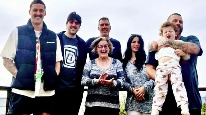 A smiling family of seven standing together outdoors, with a child being held aloft.