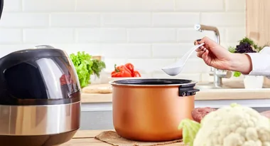 A hand holds a spoon over an open slow cooker in a kitchen with vegetables on the counter.
