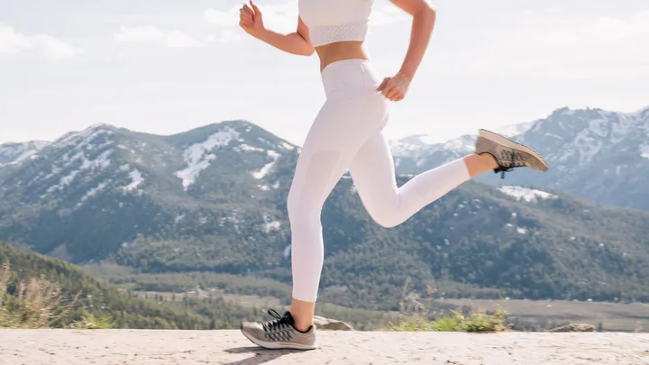 A person jogging in white leggings and a crop top, set against a scenic mountain backdrop.