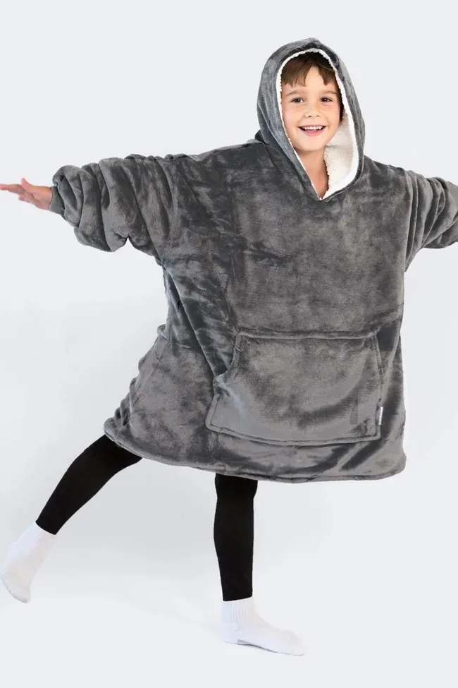 Child wearing oversized grey hooded fleece blanket with arms outstretched, smiling in white socks.