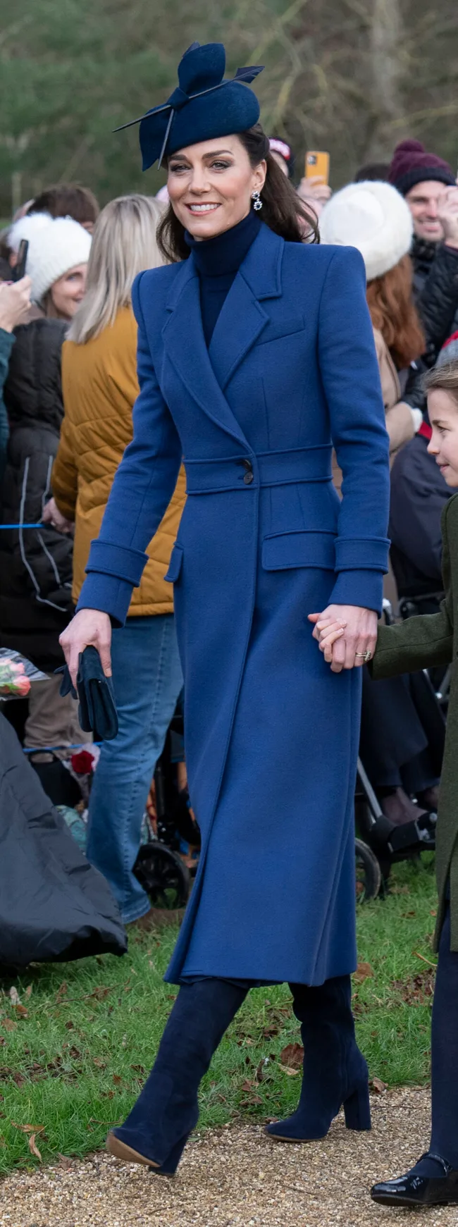 Elegant woman in a blue coat and matching hat, smiling while walking outside, holding a child's hand.