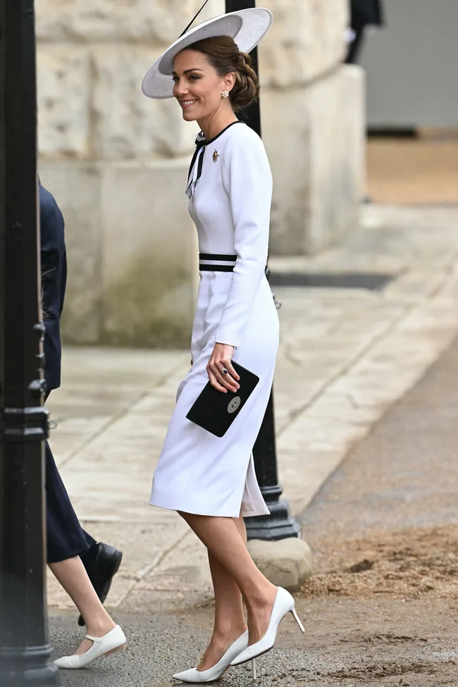 Kate Middleton in a white dress with black accents and a matching hat, holding a black clutch, smiling outdoors.