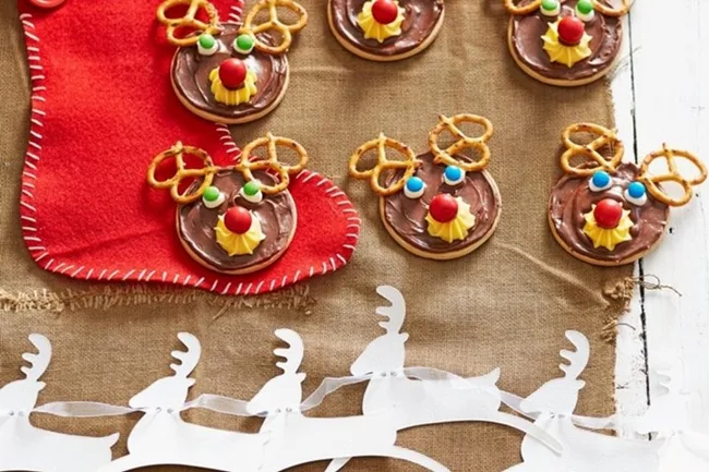 Reindeer cookies with pretzel antlers and candy faces, next to red stocking on burlap, festive decoration.