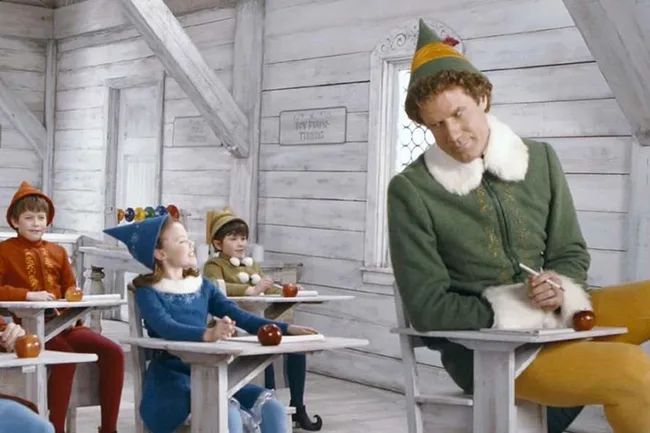 Buddy the Elf in oversized desk at school with other elves, from the movie "Elf". Room has white walls and apples on desks.