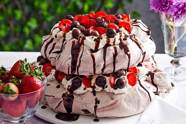Double-layered chocolate pavlova with whipped cream, strawberries, cherries, and chocolate drizzle, beside a bowl of strawberries.