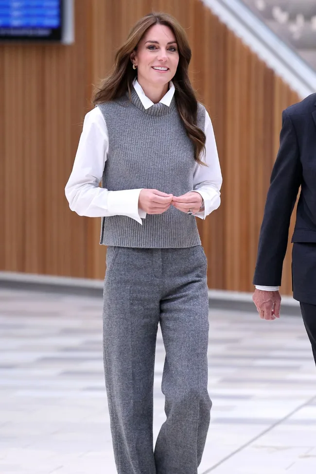 A woman in a grey knit vest and trousers over a white shirt, walking indoors with a smile.