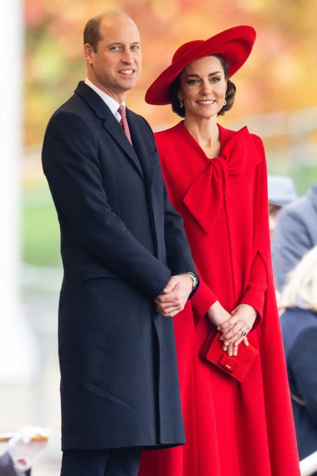 A couple stands outdoors; the woman wears a red dress and hat, while the man wears a dark coat, smiling in a formal setting.