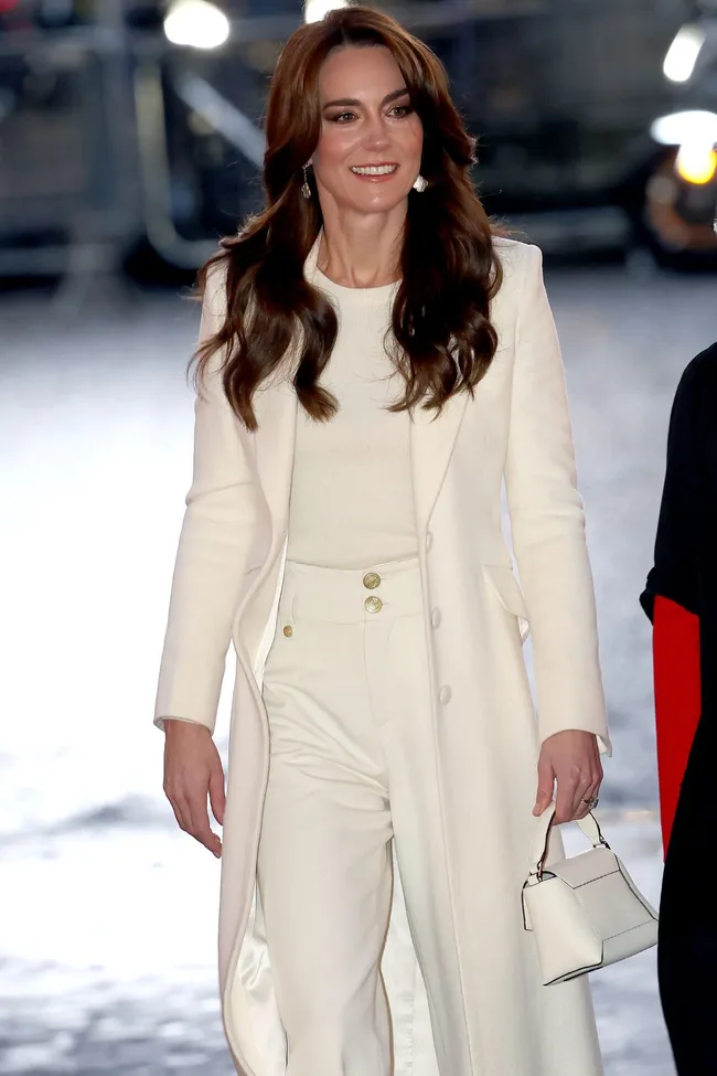 Kate Middleton in an elegant white coat and pants, holding a small white handbag, smiling.