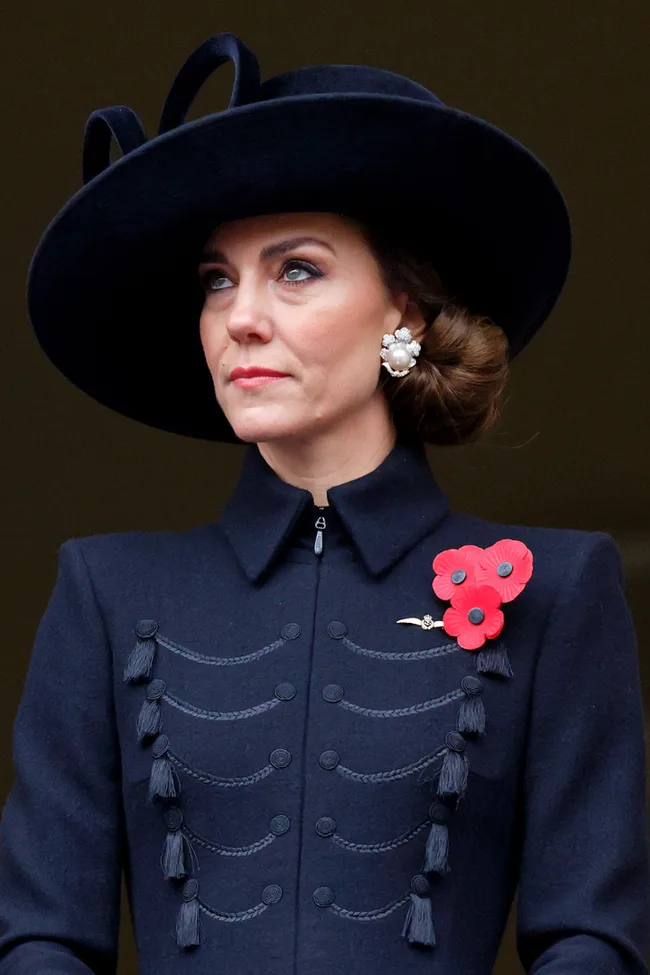 A woman in a dark coat and wide-brimmed hat with poppy brooches and pearl earrings.