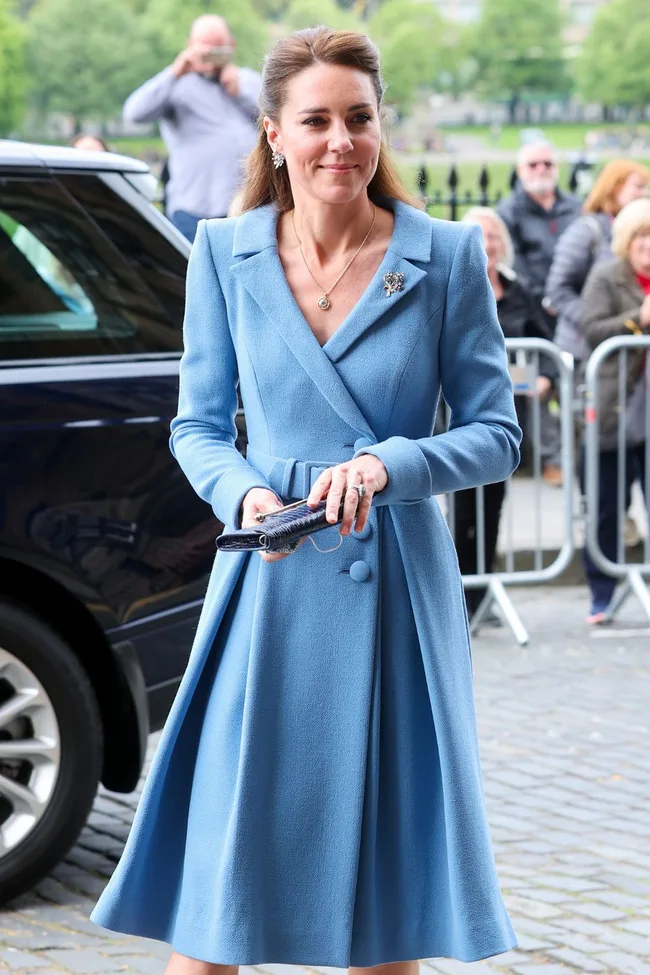 A woman in a blue coat dress stands outside a vehicle, holding a clutch, with people and trees in the background.