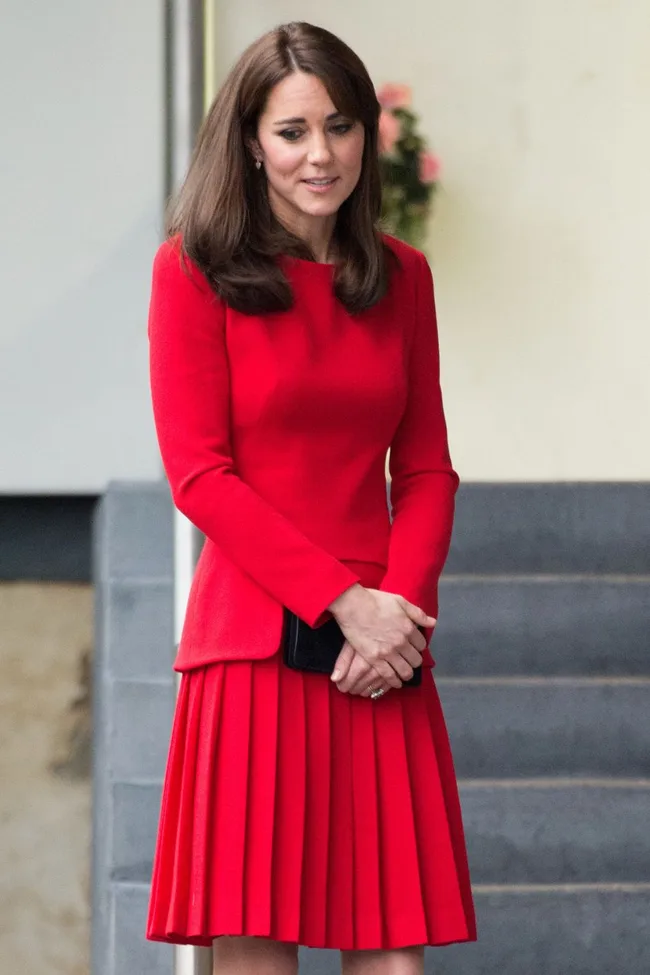 A woman in a red dress stands with her hands clasped, looking down, in front of gray stairs.