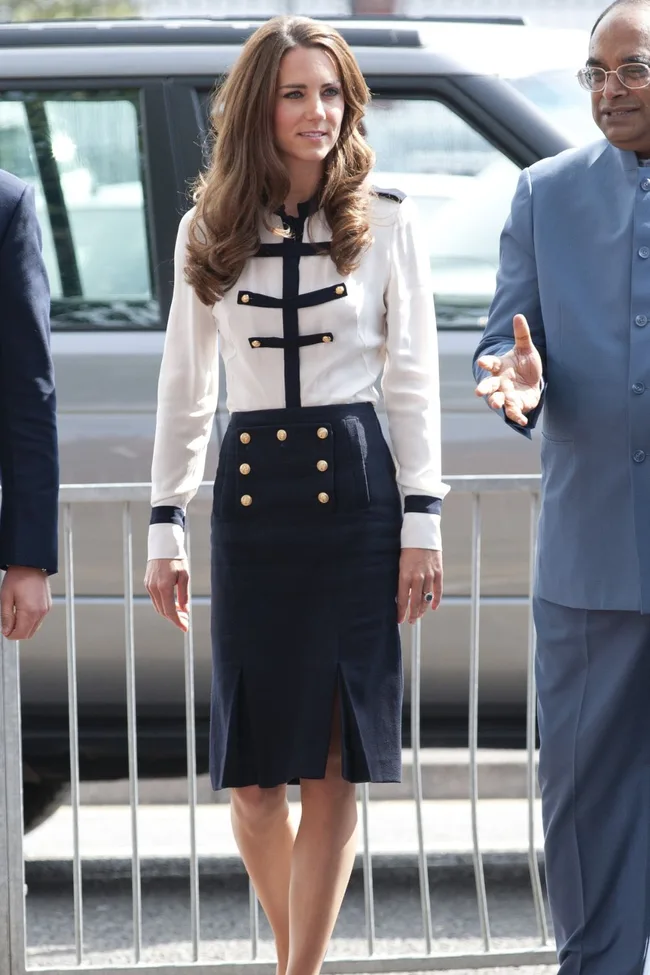 A woman in a white and navy outfit stands next to a person in a blue suit, outdoors in front of a car.