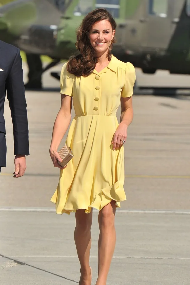 A woman in a yellow dress walks on a tarmac, holding a clutch, with a helicopter in the background.