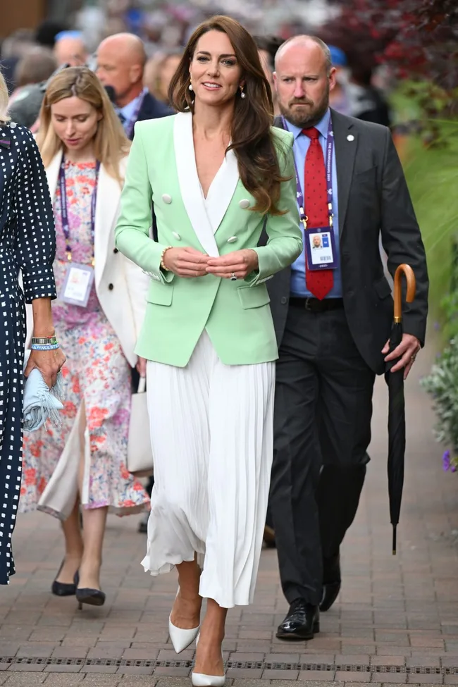 Elegant woman in a mint-green blazer and white dress walking outdoors, surrounded by people.