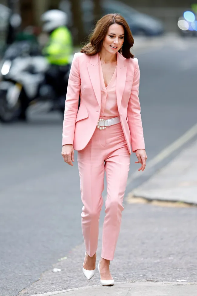 A woman in a pink suit walks on the street, wearing white heels, with a motorbike and police in the background.