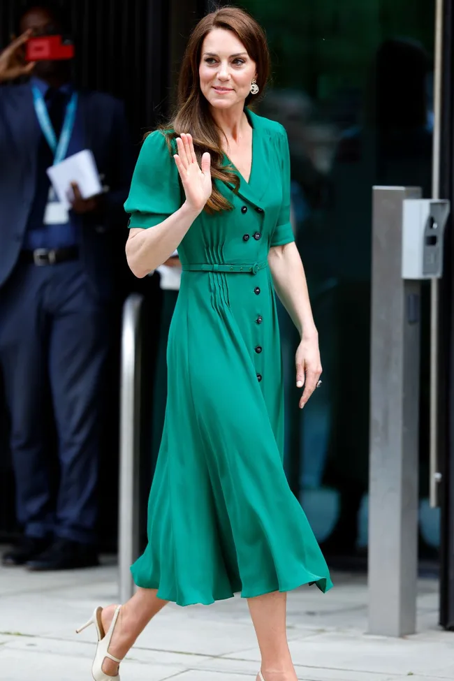 Woman in a green dress waves outside a building, smiling politely.