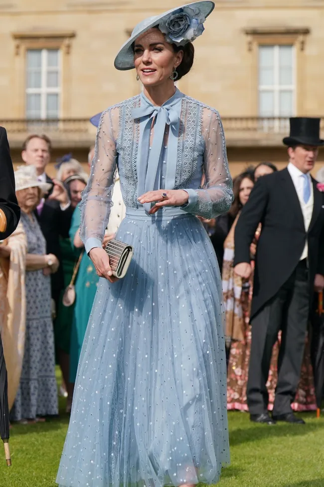 Woman in elegant blue dress with matching hat and clutch at outdoor event.