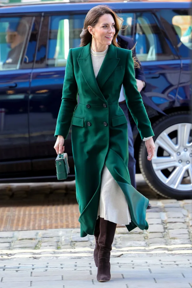 Woman in a stylish green coat and brown boots walking on a cobblestone path, holding a small green handbag.