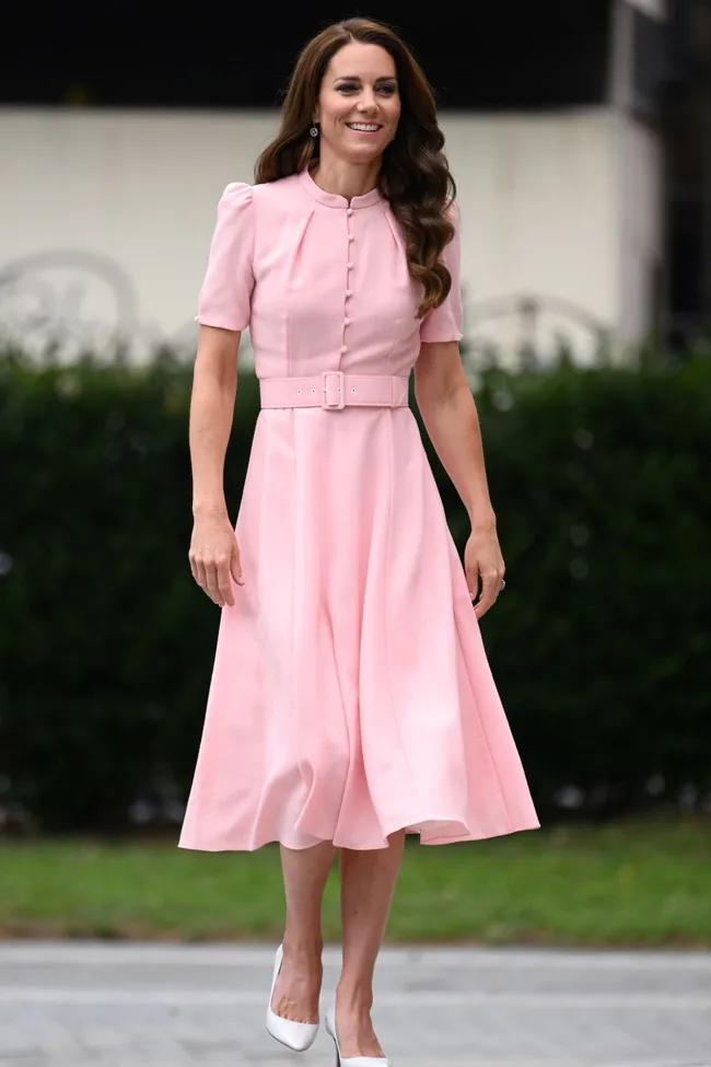 A woman wearing a pink knee-length dress with a belt and white heels, standing outside on a sidewalk.