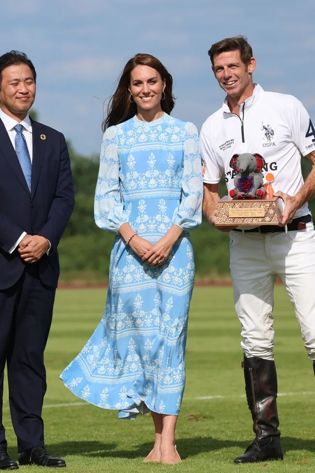 Three people standing on a grassy field, one holding a trophy with an elephant plush on it; middle person in blue dress.
