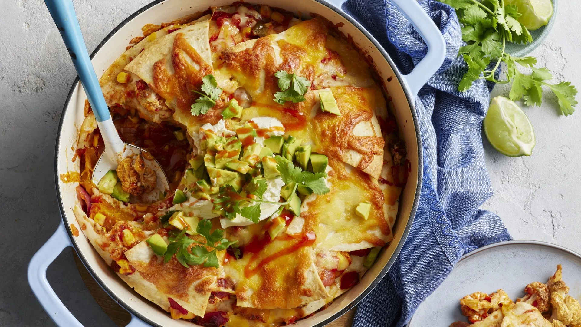 Baked enchilada skillet topped with avocado, cilantro, sour cream, and hot sauce, with serving spoon.