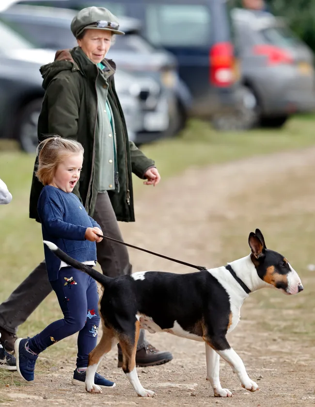 A child walking a black and white dog on a leash alongside an older woman in a green coat and cap.