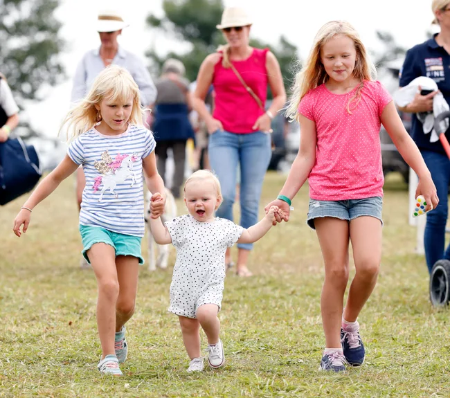 Children walking hand-in-hand on grass, smiling, with adults in the background.