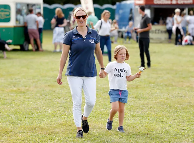 Woman in sunglasses and child holding an ice cream cone walking on grass at an outdoor event.