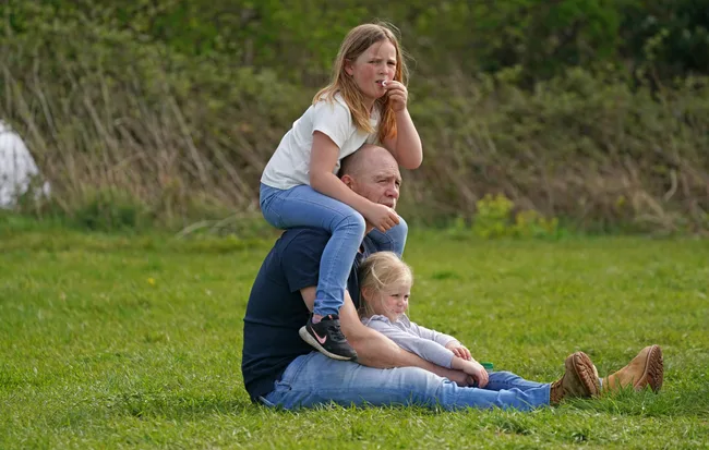 Man sitting on grass with child on shoulders and another child in his lap, set against a green natural background.