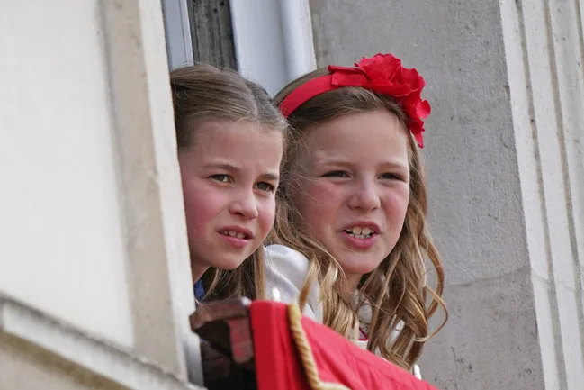 Two young girls with long hair lean out of a window, one wearing a red headband with a flower, both looking intently.