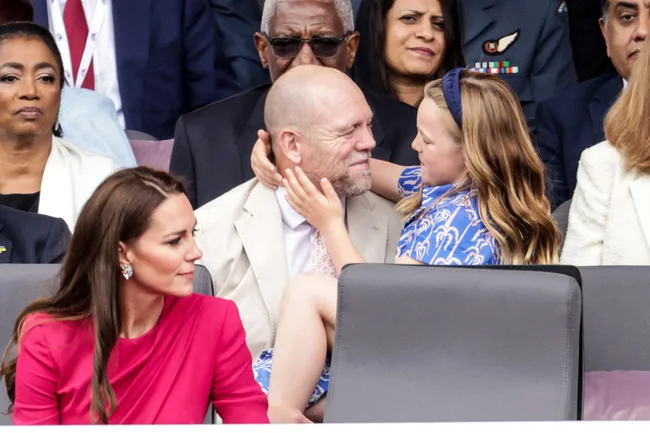 A young girl embraces a smiling bald man in a crowded event, while a woman in a pink dress looks on.