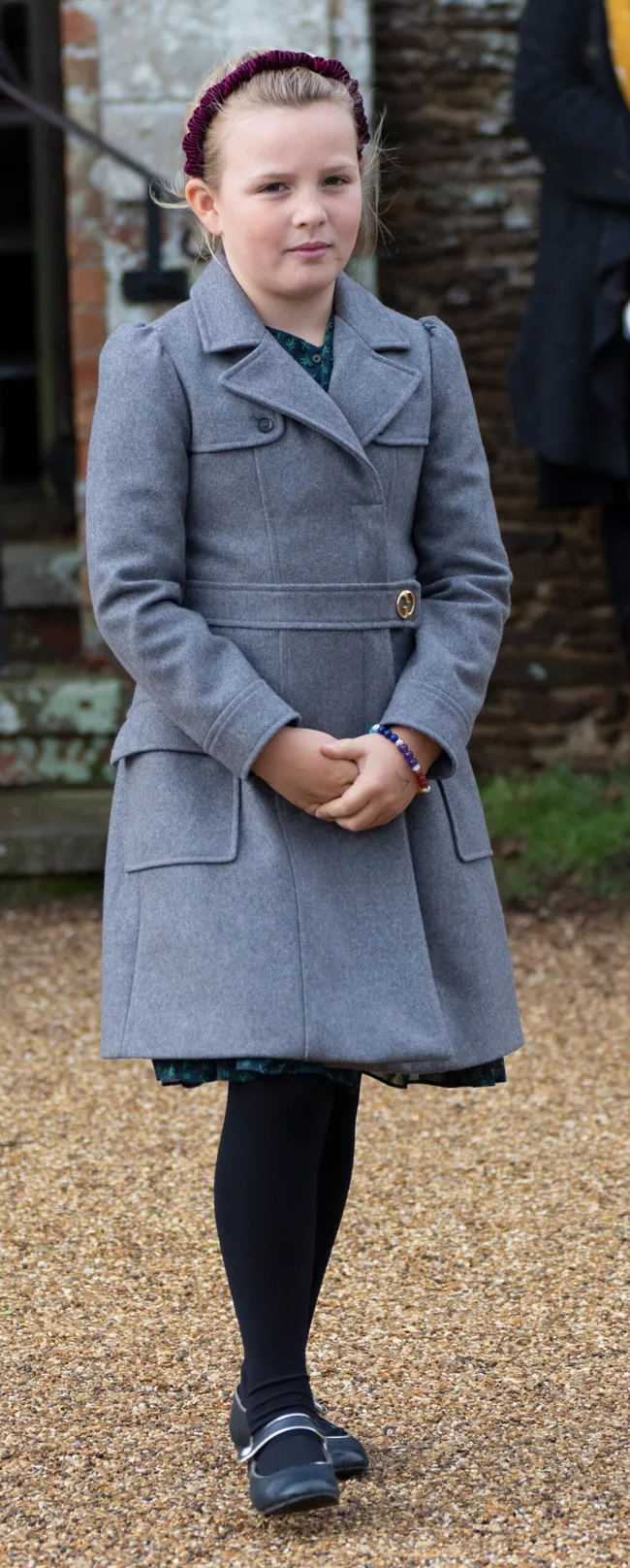 Child in grey coat with maroon headband, standing on gravel path outside a brick building.