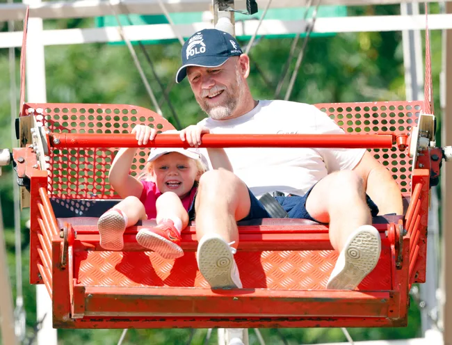 A smiling man and young child enjoy a ride on a red Ferris wheel seat, with trees visible in the background.