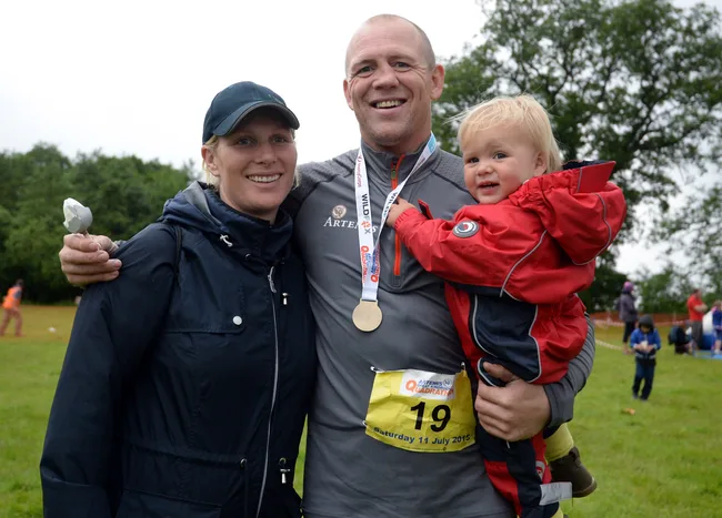 Family outdoors, smiling. Man with medal, child in red, woman in cap. Event with trees and people in background.