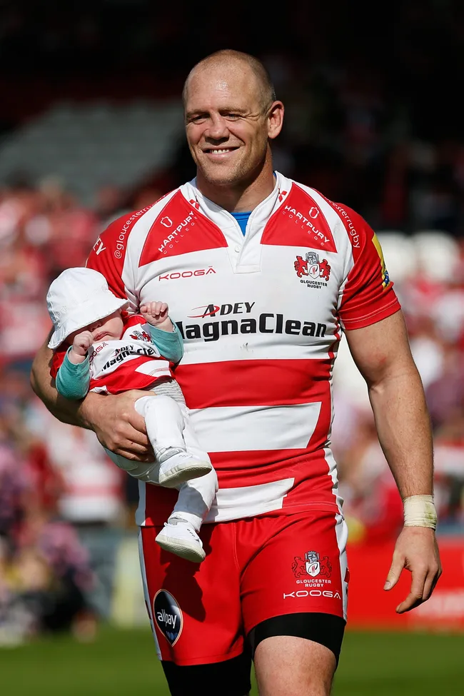 Rugby player in a red and white striped jersey carrying a baby dressed similarly on a sports field.
