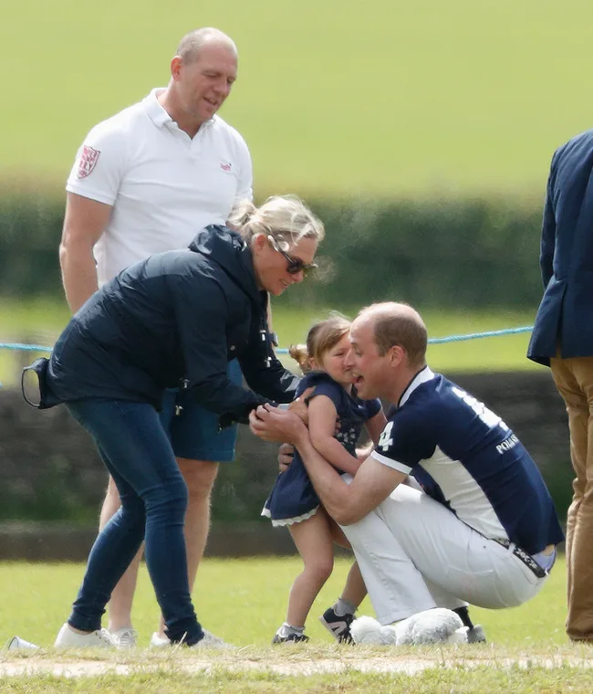 People gathered outdoors, one man kneels to comfort a child, surrounded by adults in casual attire, grassy field background.