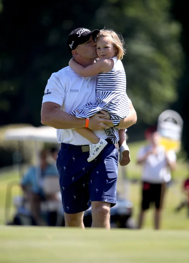 Man in a golf outfit holding and kissing a young girl in a striped dress outdoors.