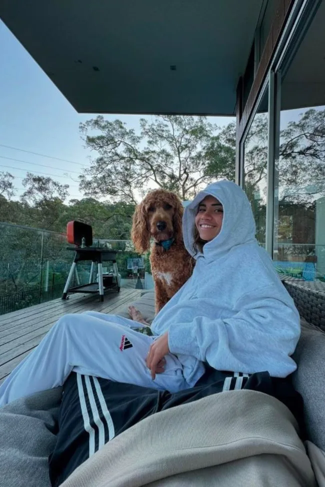 Person in a hoodie relaxing on a patio with a brown dog, trees in the background.
