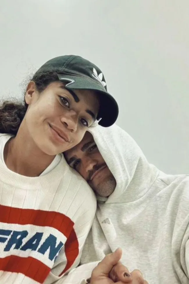 Couple smiling; woman wearing a cap, man in hoodie, holding hands.