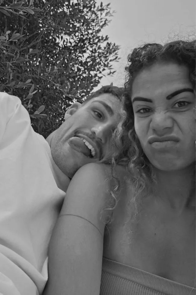 Two people making playful faces near foliage, one with tongue out, the other with furrowed brow, in black and white.