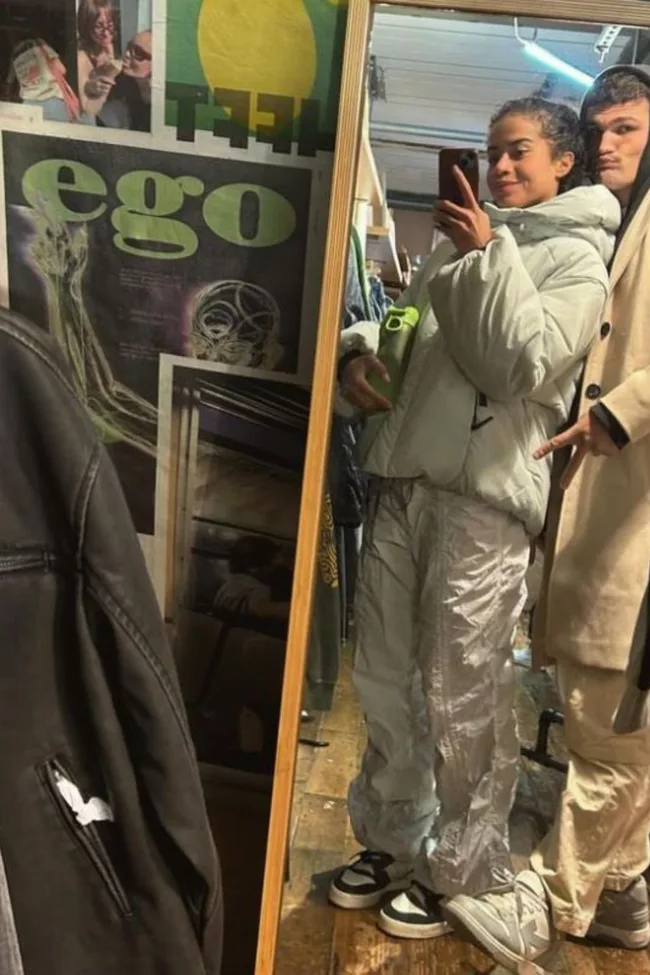 Two people taking a mirror selfie; one in a white puffer jacket, holding a green bottle. Posters are visible in the background.