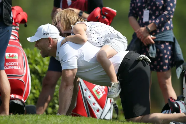 Man giving piggyback ride to child at a golf event, with golf bags in the background.