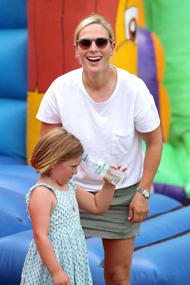 A woman with sunglasses and a young girl holding a water bottle stand smiling in front of colorful inflatable equipment.